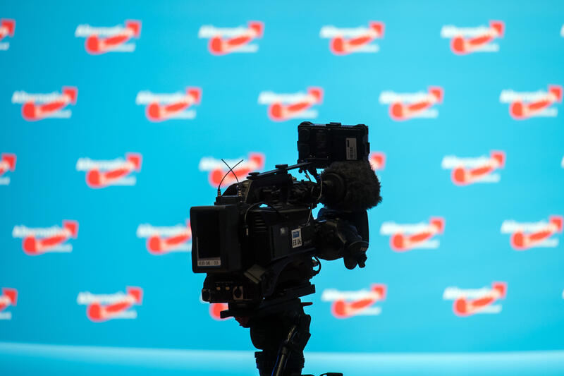 s:27:"Fernsehkamera vor AfD-Logos";
