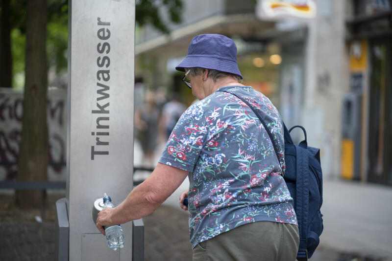 s:50:"Seniorin erfrischt sich an einer Trinkwassersäule";
