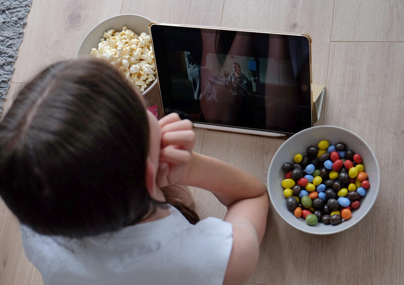 s:68:"Ein Mädchen schaut eine Serie auf ihrem iPad und isst Süssigkeiten";
